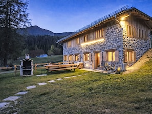 Ein Steinhaus mit einer Holzterrasse, einem Grill und einem Berg im Hintergrund.