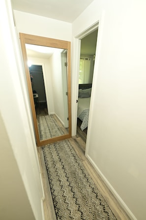 A hallway with a wooden-framed mirror, a patterned rug, and a bed with a checkered bedspread.
