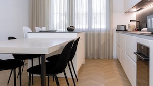 Une cuisine moderne avec un coin repas, dotée d’un parquet en chevrons, de luminaires suspendus et d’une grande fenêtre agrémentée de rideaux légers.