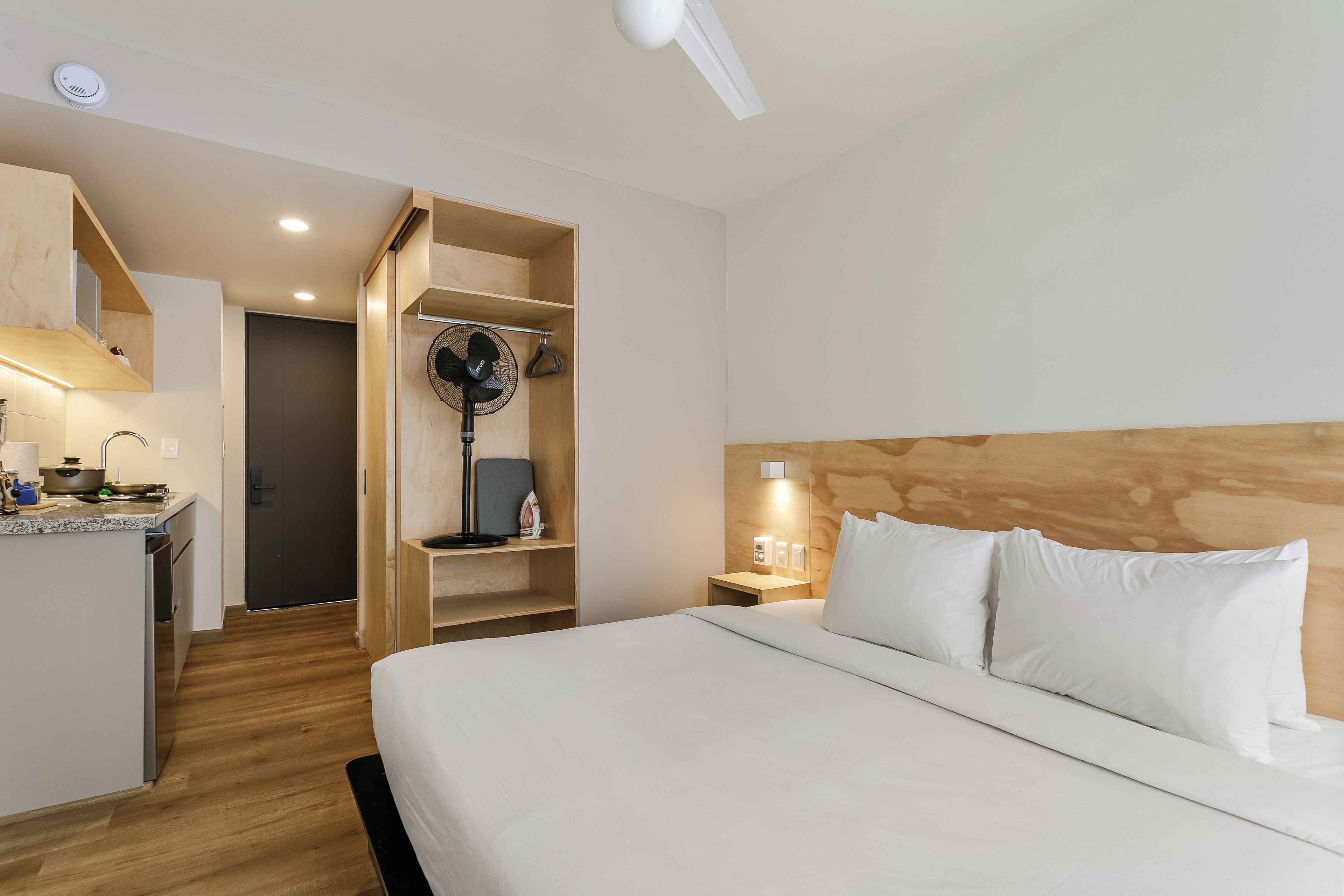 A hotel room with a bed, bedside table, and a kitchenette area.