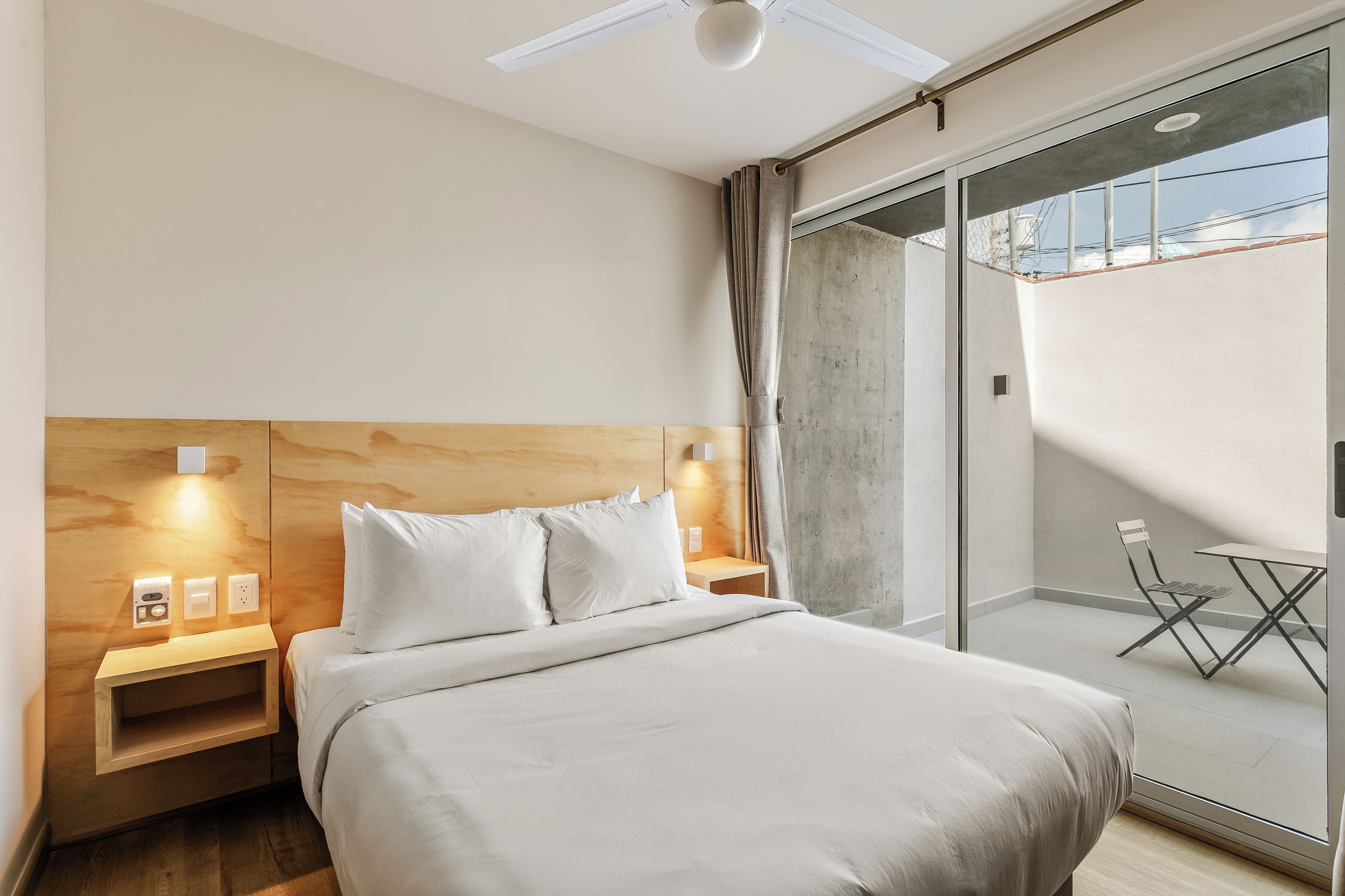 A modern hotel room with a bed, bedside tables, a ceiling fan, and a view of the outside.