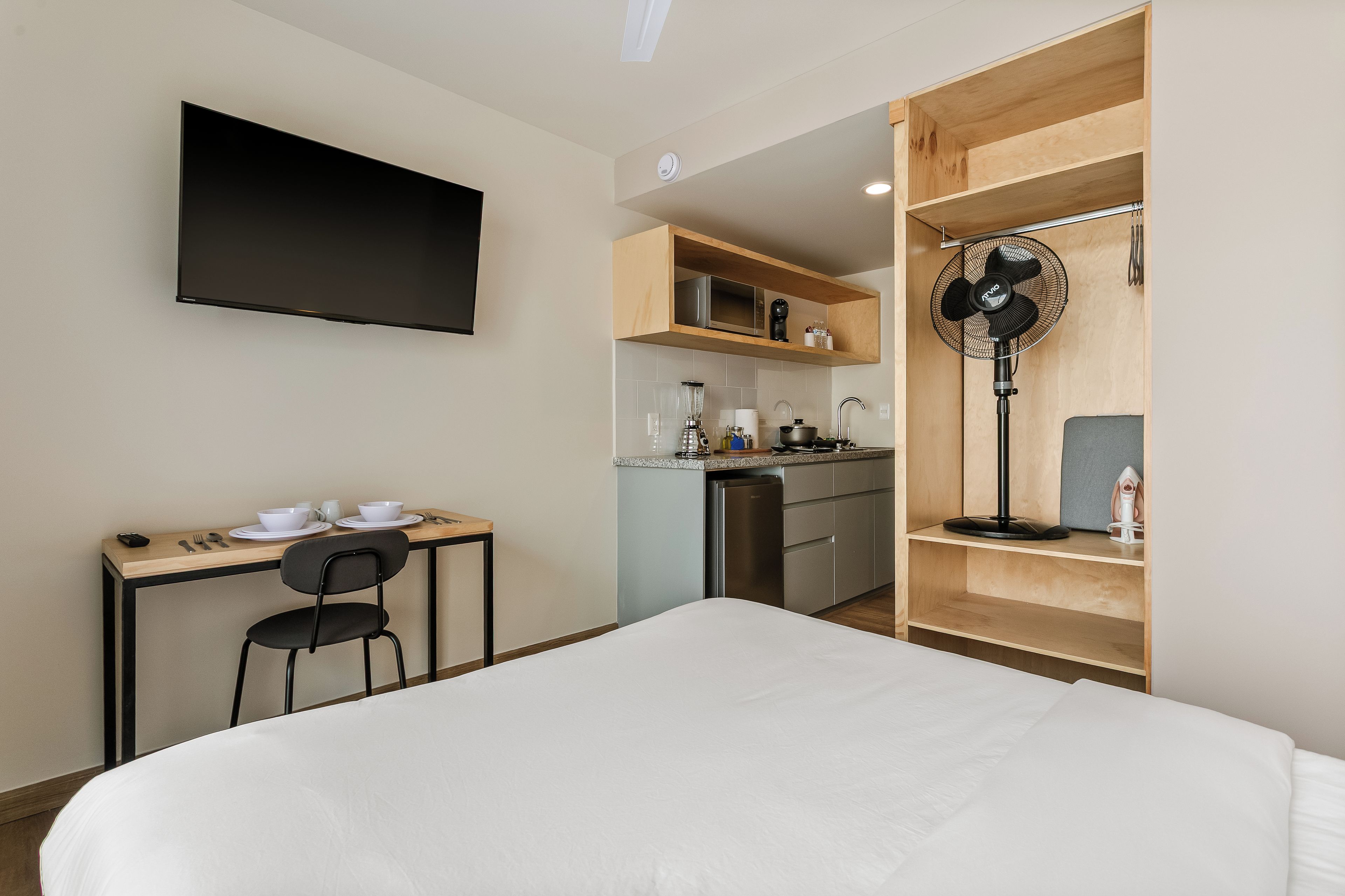 Habitación de hotel compacta con cama, kitchenette pequeña, mesa de comedor con sillas y televisor montado en la pared.