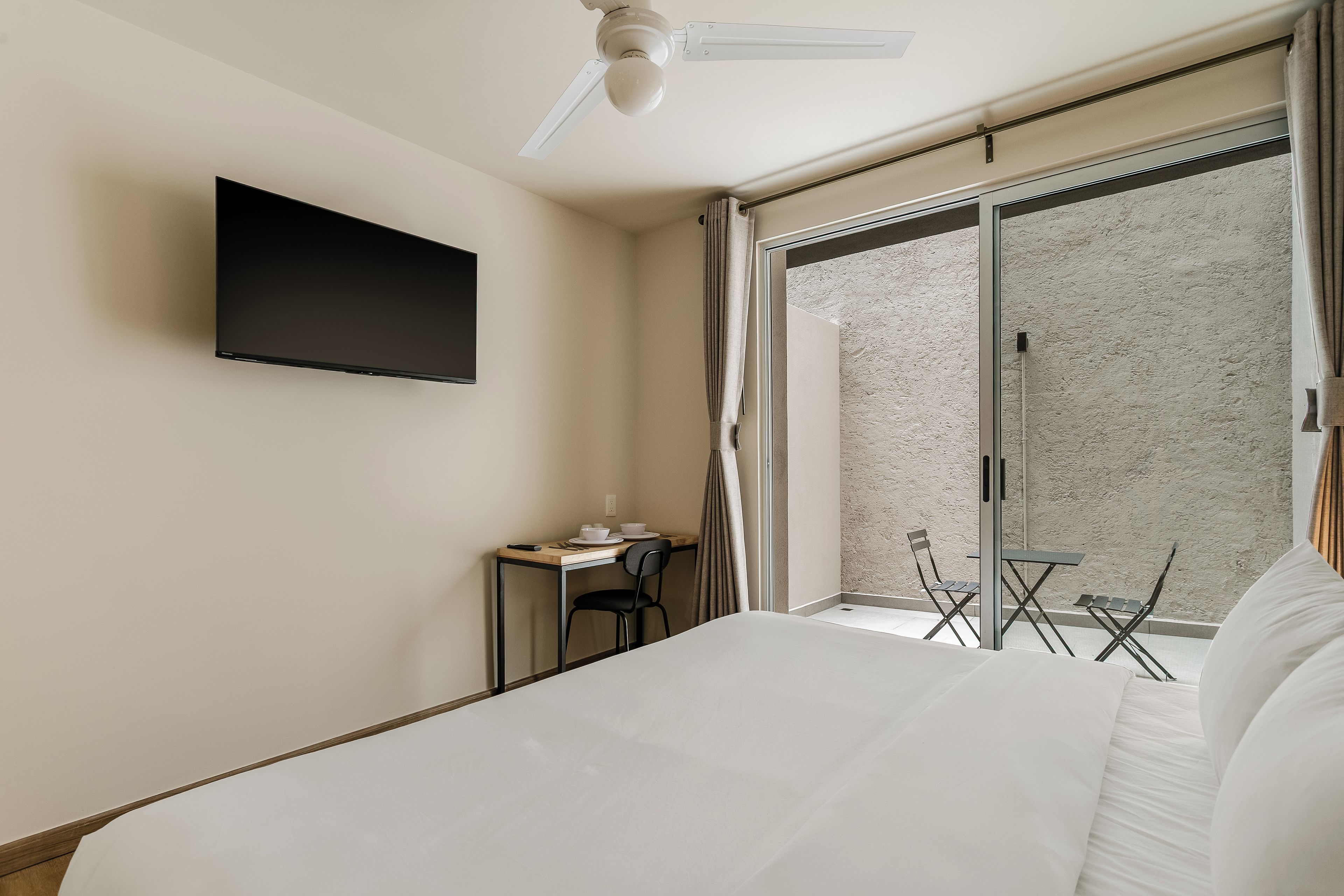 Habitación de hotel con cama, escritorio con silla, televisor montado en la pared y puerta corrediza de vidrio que da a un área exterior.