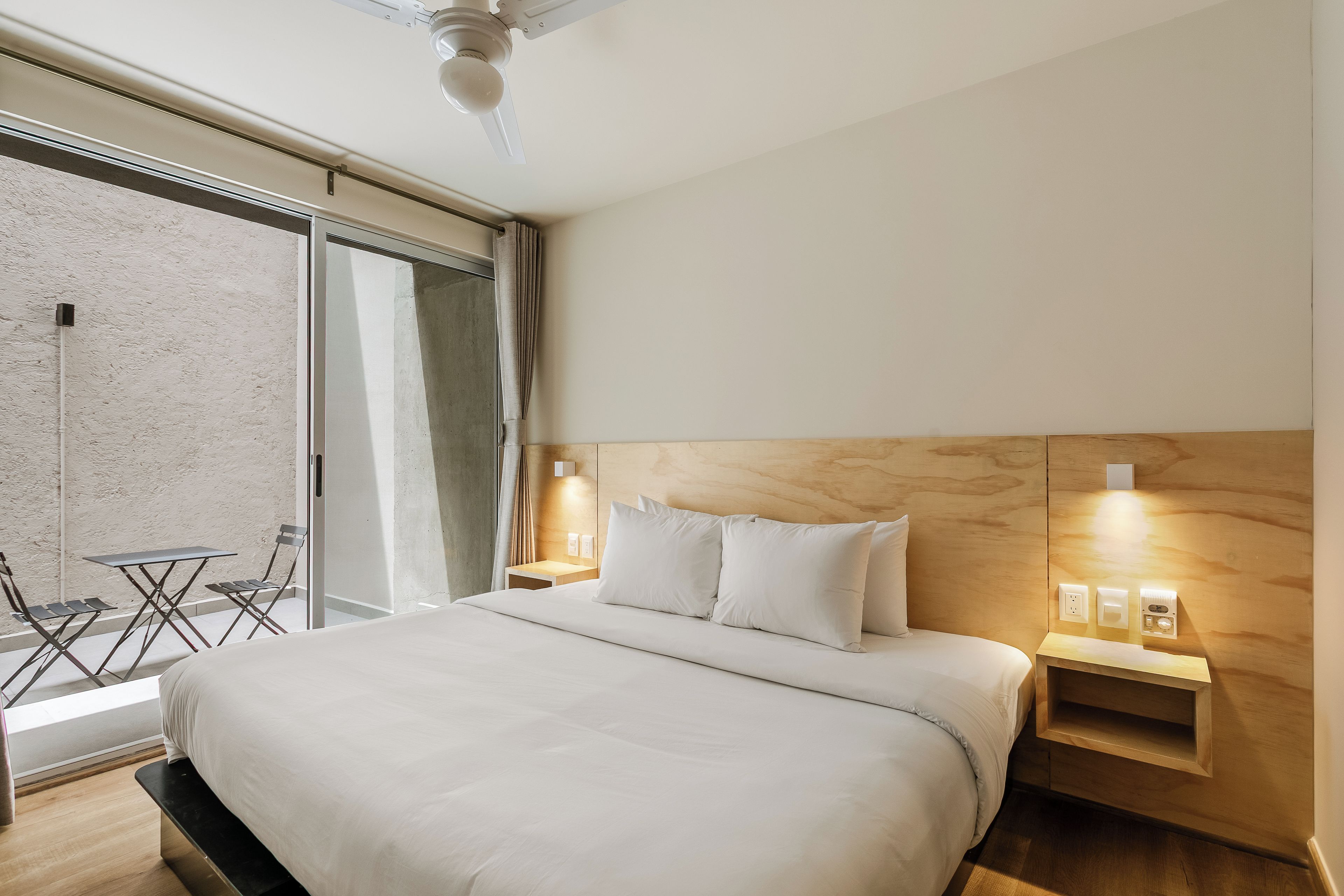 A hotel room with a bed, a nightstand, a wall-mounted light, and a view of the outside.
