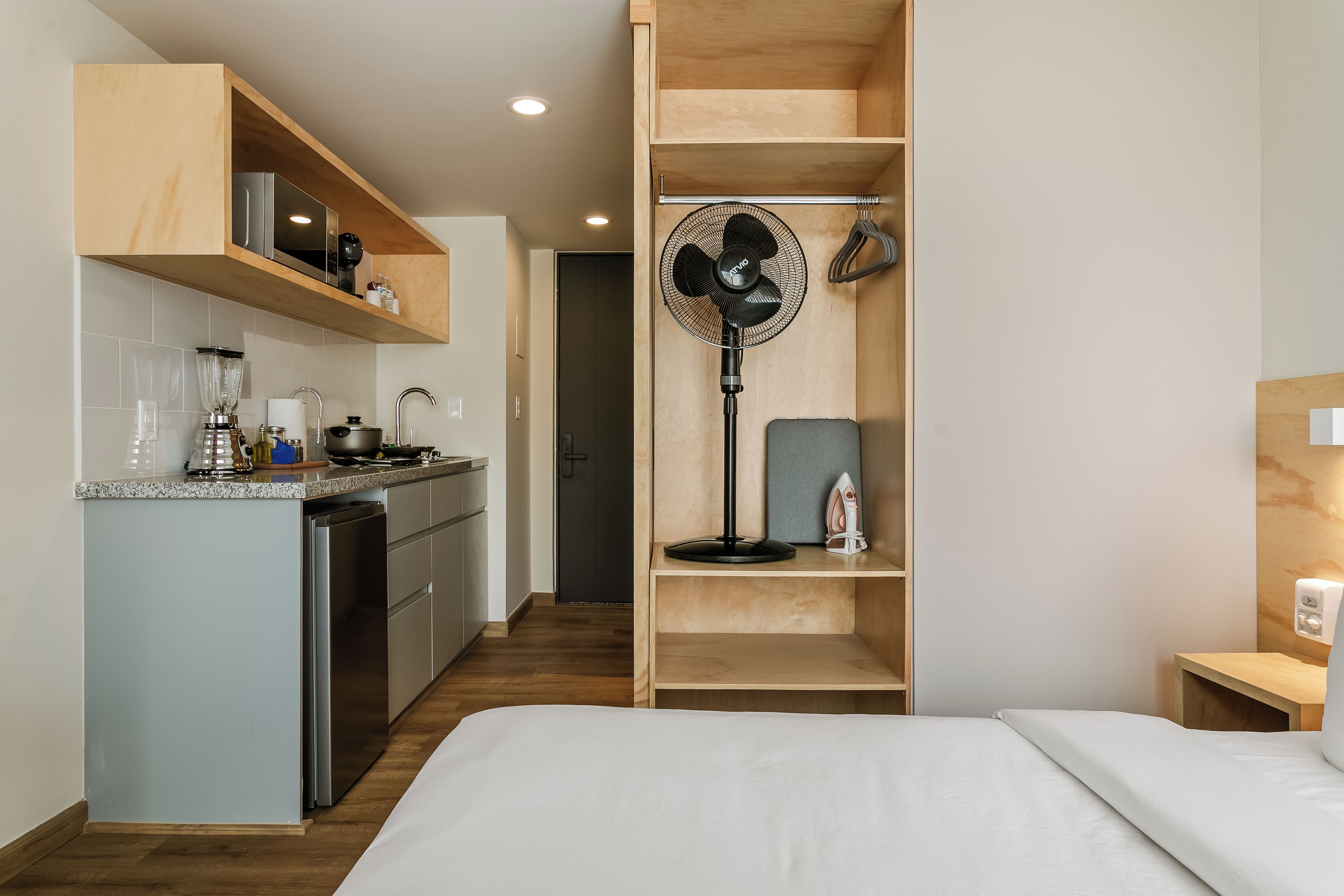 A compact hotel room with a kitchenette, a bed, a standing fan, and a small ironing station.