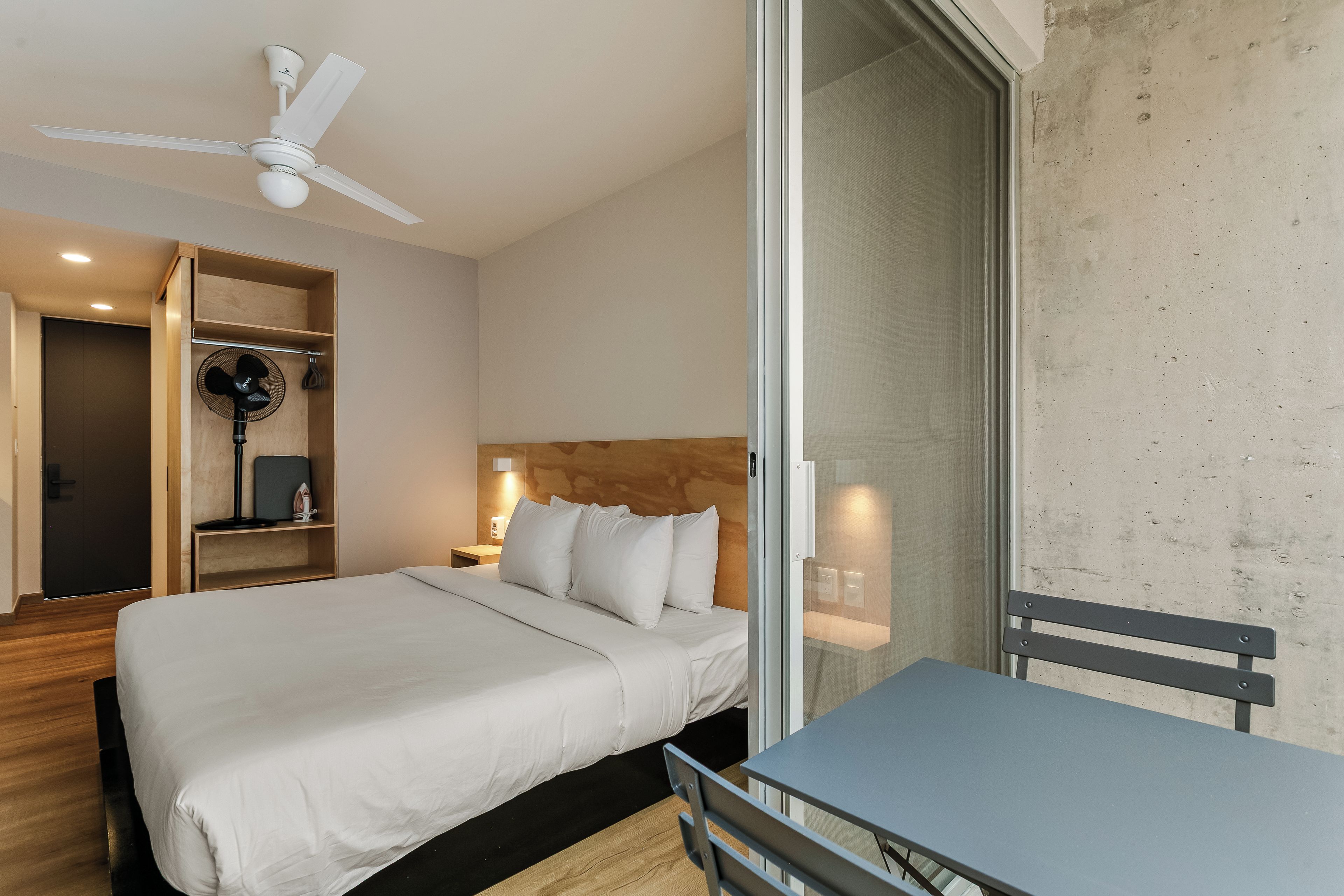 A hotel room with a bed, a table, a ceiling fan, and a view of the outside.