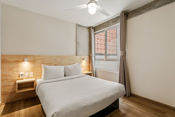 Un dormitorio con cabecera de madera, cama con ropa de cama blanca, mesita de noche con lámpara y ventana con cortinas.
