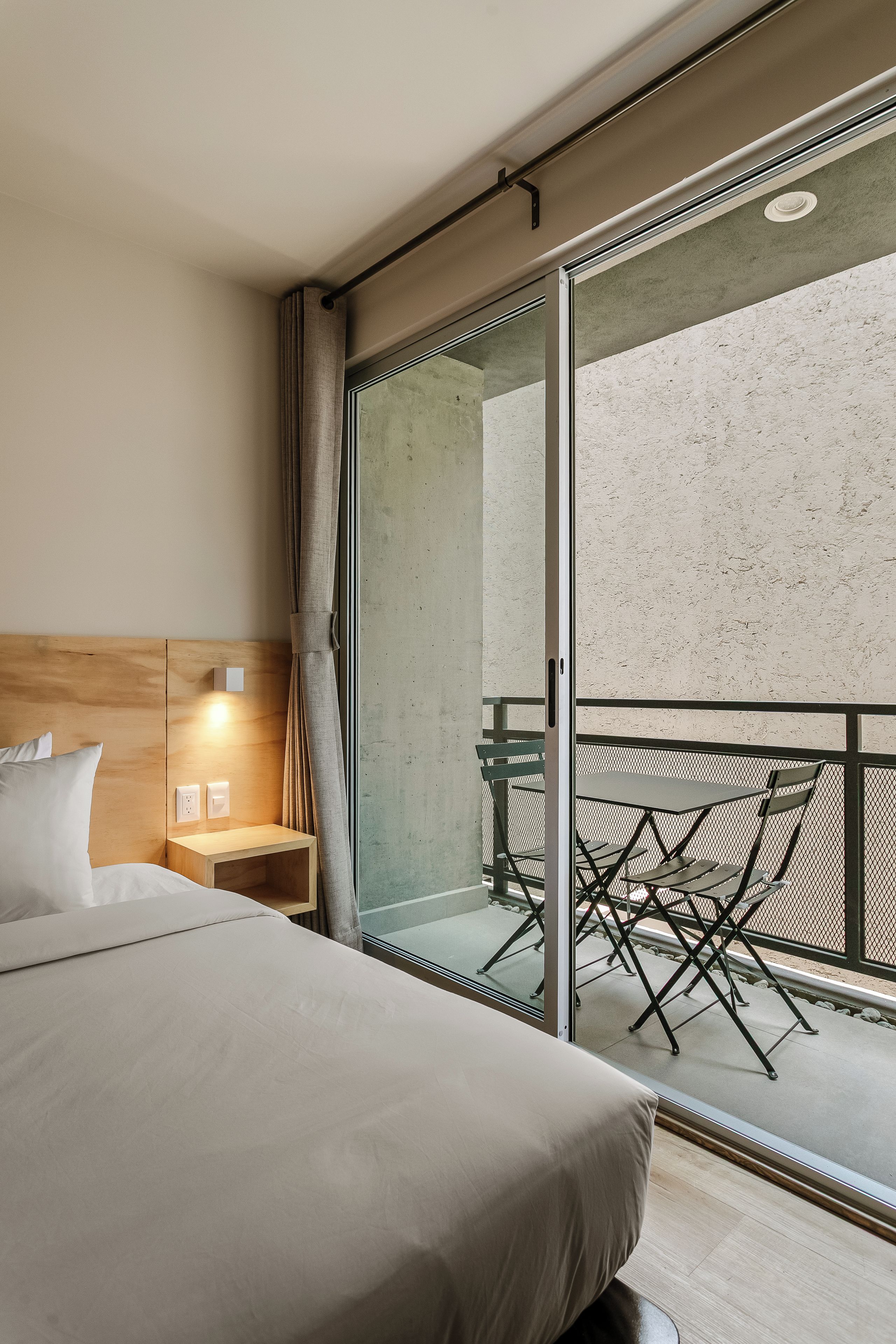 Habitación de hotel con cama, mesita de noche, lámpara y vistas a un balcón con mobiliario exterior.