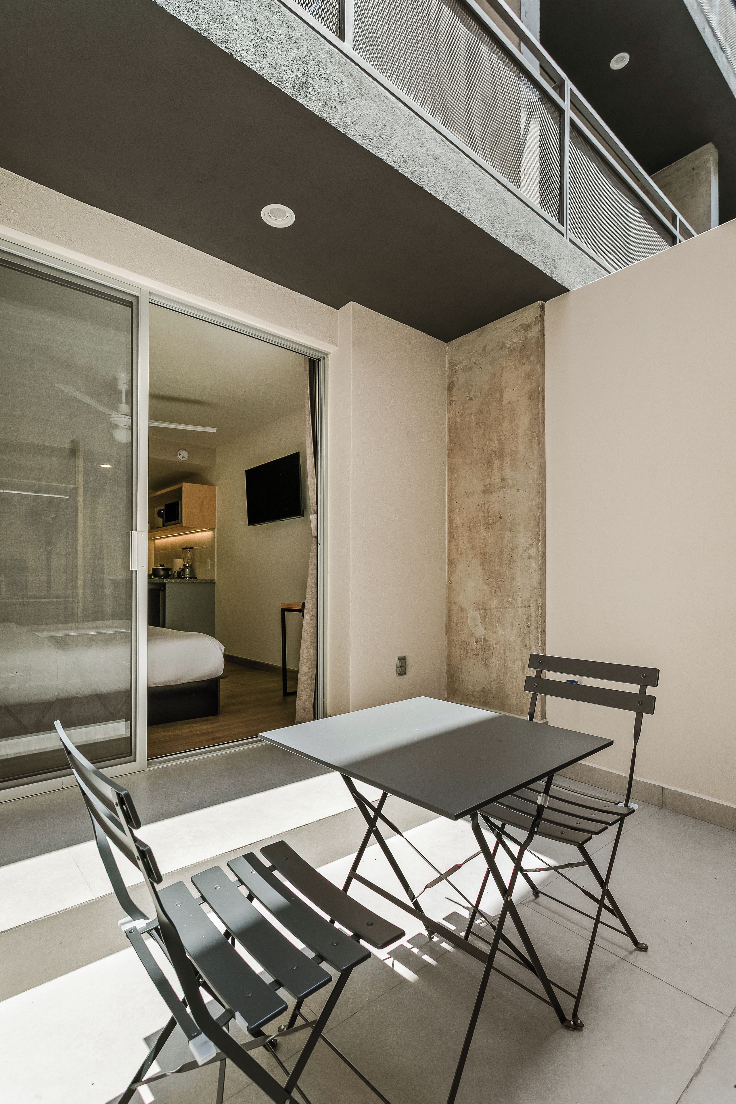 Un patio exterior moderno con una mesa y sillas, un ventanal corredero que da a un dormitorio y una cocina al fondo.