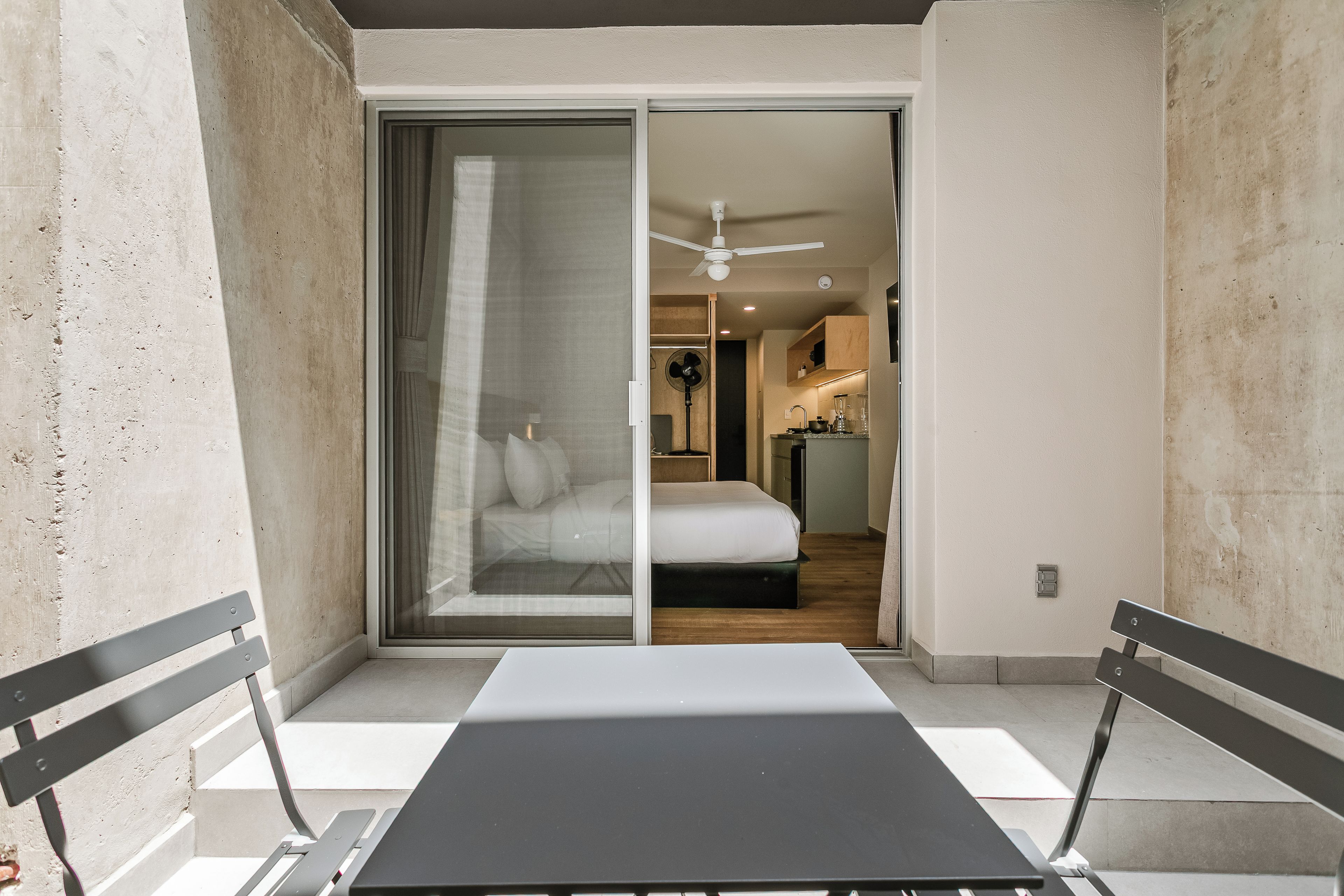 Habitación de hotel con puerta de vidrio corrediza que da a un balcón, una cama, una pequeña kitchenette y un ventilador de techo.