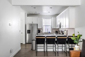 Eine moderne Küche mit weißen Schränken, einem Edelstahl-Kühlschrank, einer Bar mit Hockern und einem Fenster mit Jalousien.