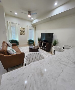 A living room with a brown sofa, a white armchair, a coffee table, a TV on a stand, and a large window with blinds. - Modern 4BR New Home - Minutes to NRG, Med. Ctr, Downtown & UH (Houston)