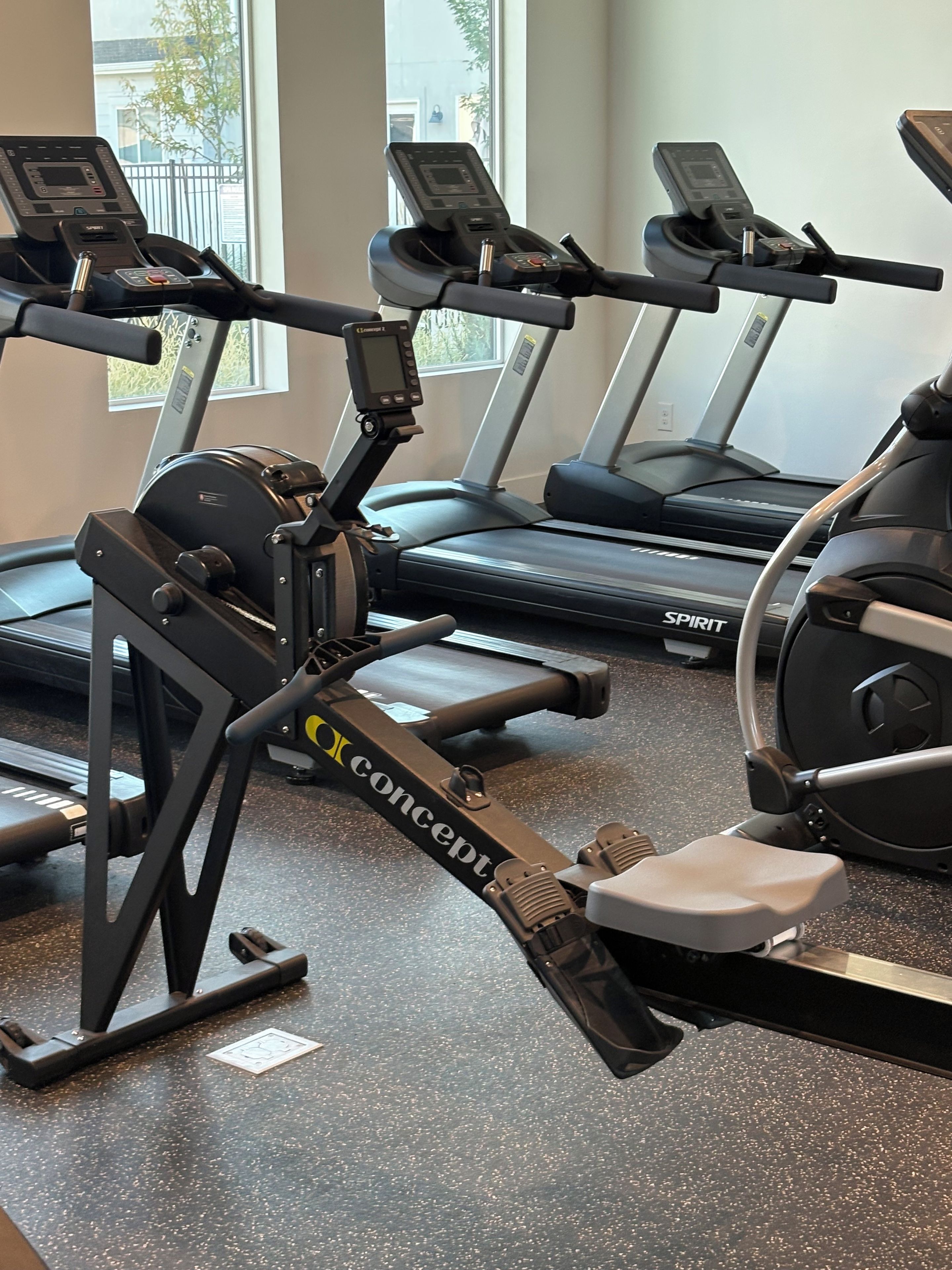 A gym with stationary bikes and treadmills, a window with a view of the outside, and a speckled floor.