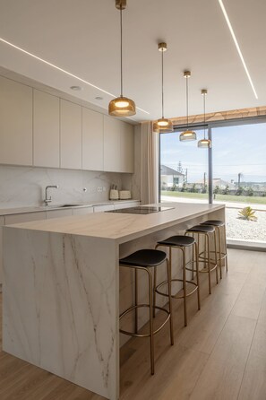 Eine moderne Küche mit Marmorarbeitsplatte, Barhocker, Pendelleuchten und Blick durch große Fenster.