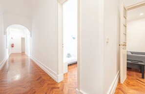 Ein Korridor mit Holzboden, weißen Wänden und einer Tür zu einem Zimmer mit einem Bett und einem grauen Sofa.