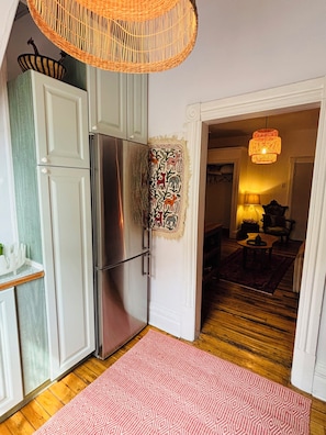 Una cocina con un refrigerador, un cesto colgante, un tapete estampado y un acceso a una sala de estar con una lámpara.