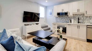 Een moderne keuken met witte kasten, een magnetron en een ingebouwde oven. Er is een flatscreen televisie aan de muur, een eettafel met stoelen en een bank met blauwe kussens.