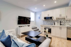 A modern kitchen with white cabinets, a microwave, and a built-in oven. There is a flat-screen TV mounted on the wall, a dining table with chairs, and a sofa with blue cushions.