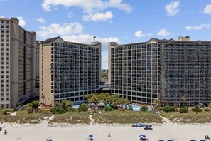 A hotel with multiple floors, a beach in the foreground, and a clear sky. - Beach Cove Resort 515 Oceanfront Condo (North Myrtle Beach)