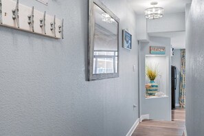 A hallway with a light gray wall, a wooden floor, a mirror, a welcome sign, and a plant in a striped vase. - Beach Cove Resort 515 Oceanfront Condo (North Myrtle Beach)