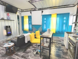 Una habitación con sofá, una silla amarilla, una mesita, televisor y una zona de cocina.