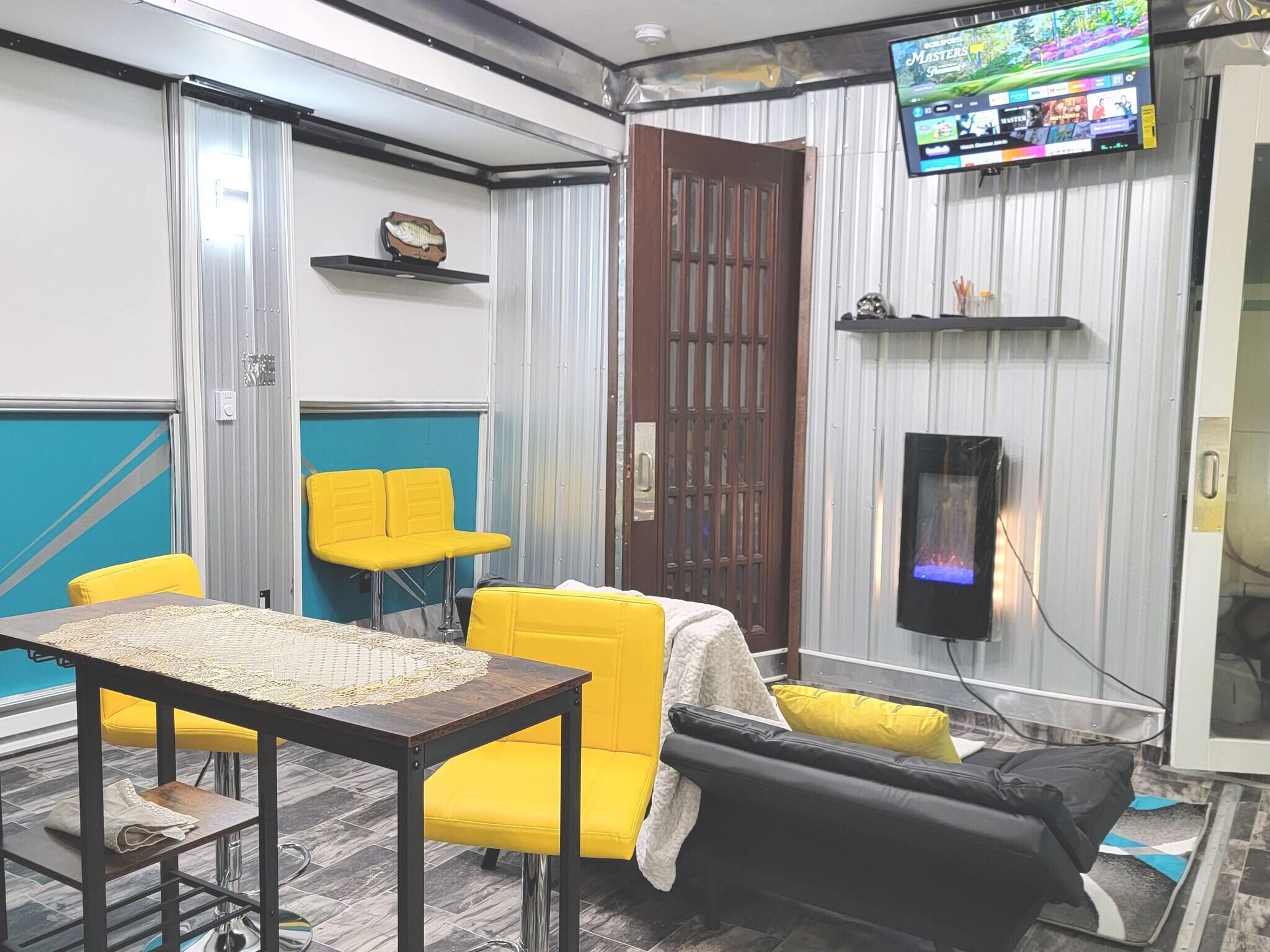 A room with a TV, yellow chairs, a table, and a black sofa.