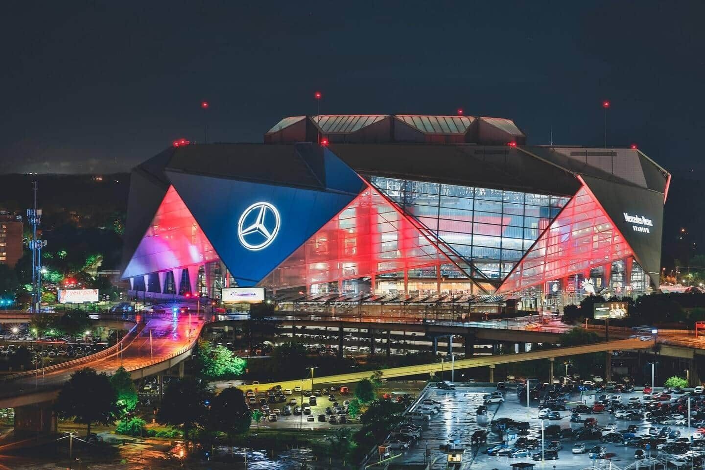 A large, modern stadium with a prominent logo on the side, surrounded by a parking area and a road.