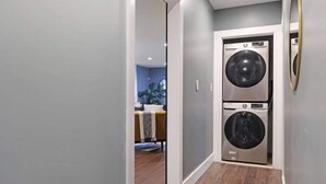 A laundry room with two stacked washing machines, a wooden floor, and a view into a living area with a sofa and a plant. - Beautiful Den 4Bd 4bth Perfect for Longer Stays (Atlanta)