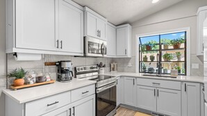 A modern kitchen with white cabinets, a built-in oven, a coffee maker, and a window with potted plants. - 6BR Boise Getaway - Arcade - Hot Tub - King Beds - Fire Pit - Sleeps 15+ (Boise)