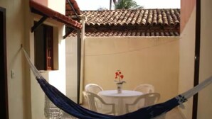 Una hamaca atada entre dos postes en un espacio exterior cubierto, con una mesa, sillas, techo de baldosas y una planta en maceta.