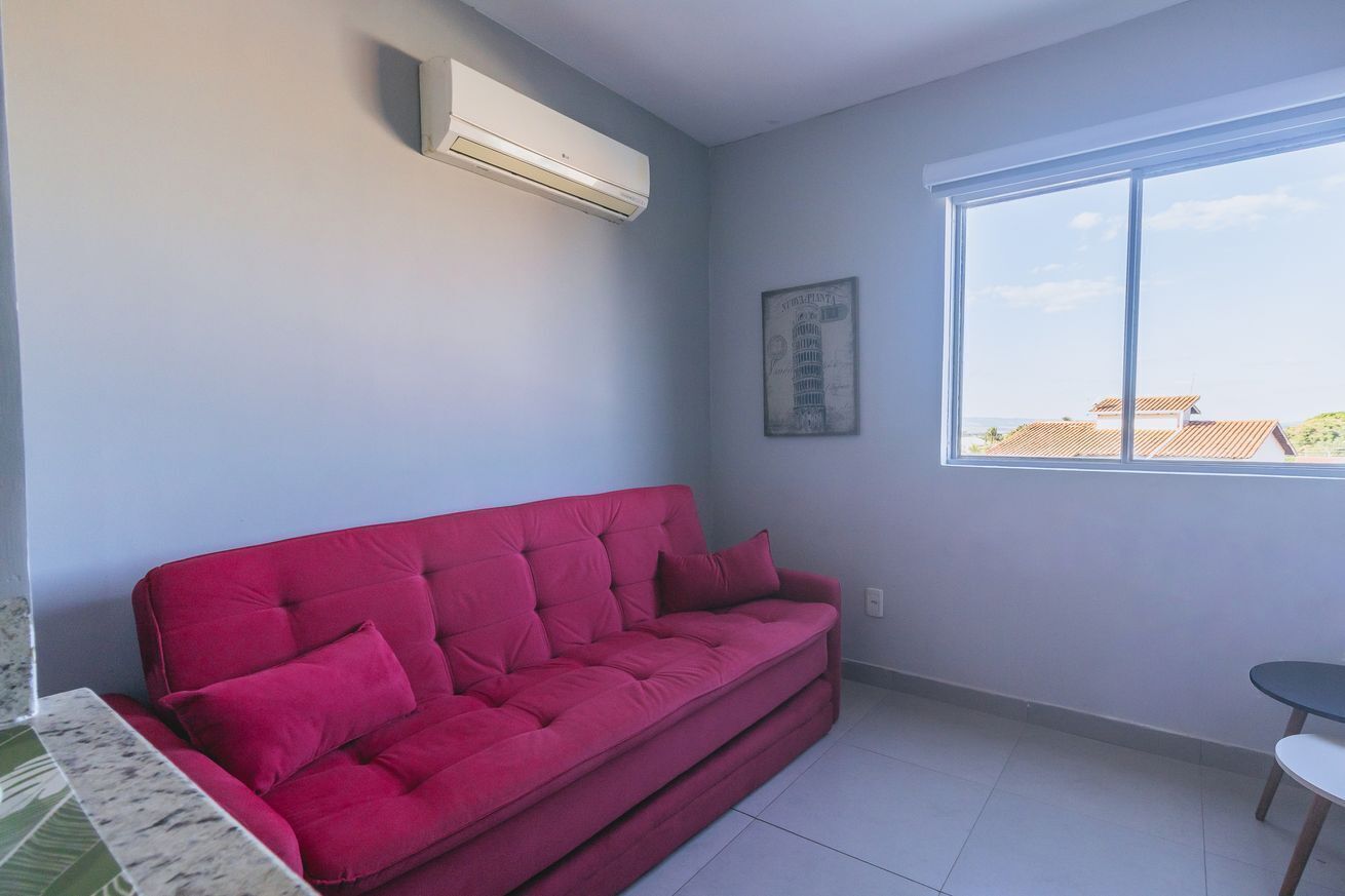 A room with a red sofa, a small table, a window with a view, and an air conditioning unit.
