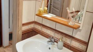Ein Badezimmer mit weißem Waschbecken, einem Spiegel, einem Holzregal mit Pflegeprodukten und einer gefliesten Wand mit dekorativen Mustern.