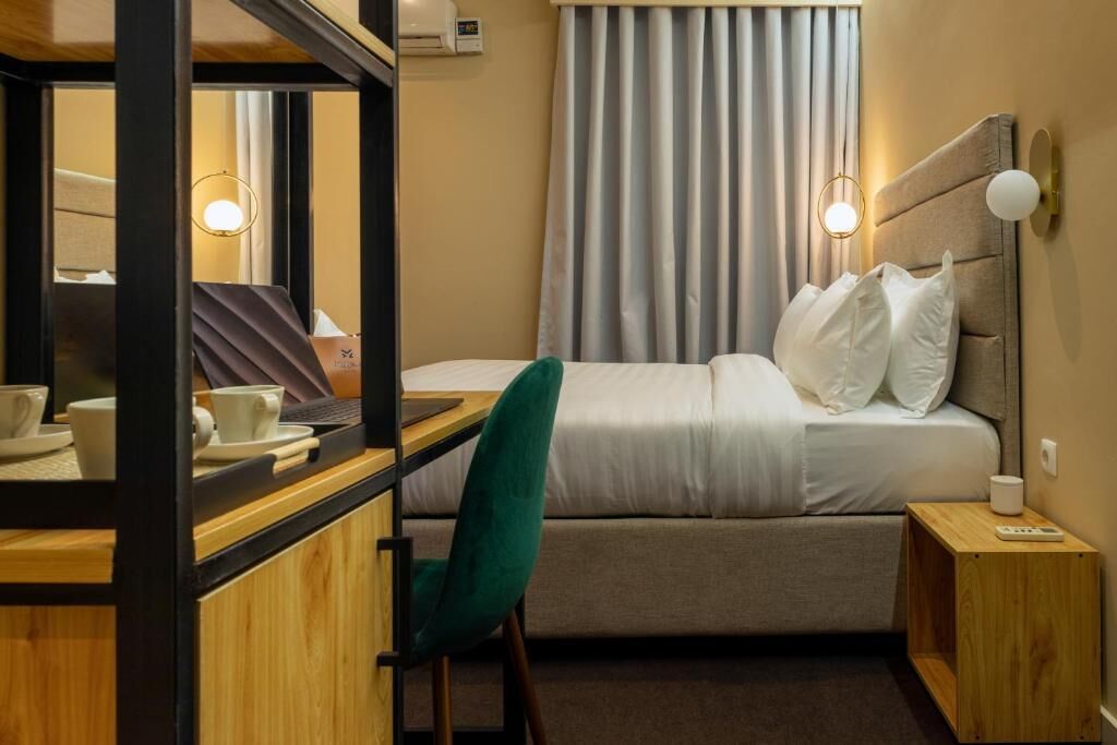 A hotel room with a bed, a desk with a chair, a mirror, a bedside table, and a window with curtains.