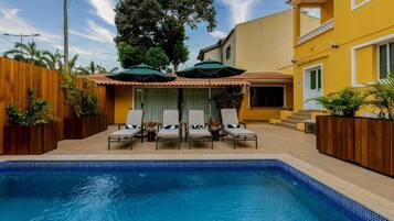 A swimming pool with lounge chairs and umbrellas, surrounded by wooden fences and plants.