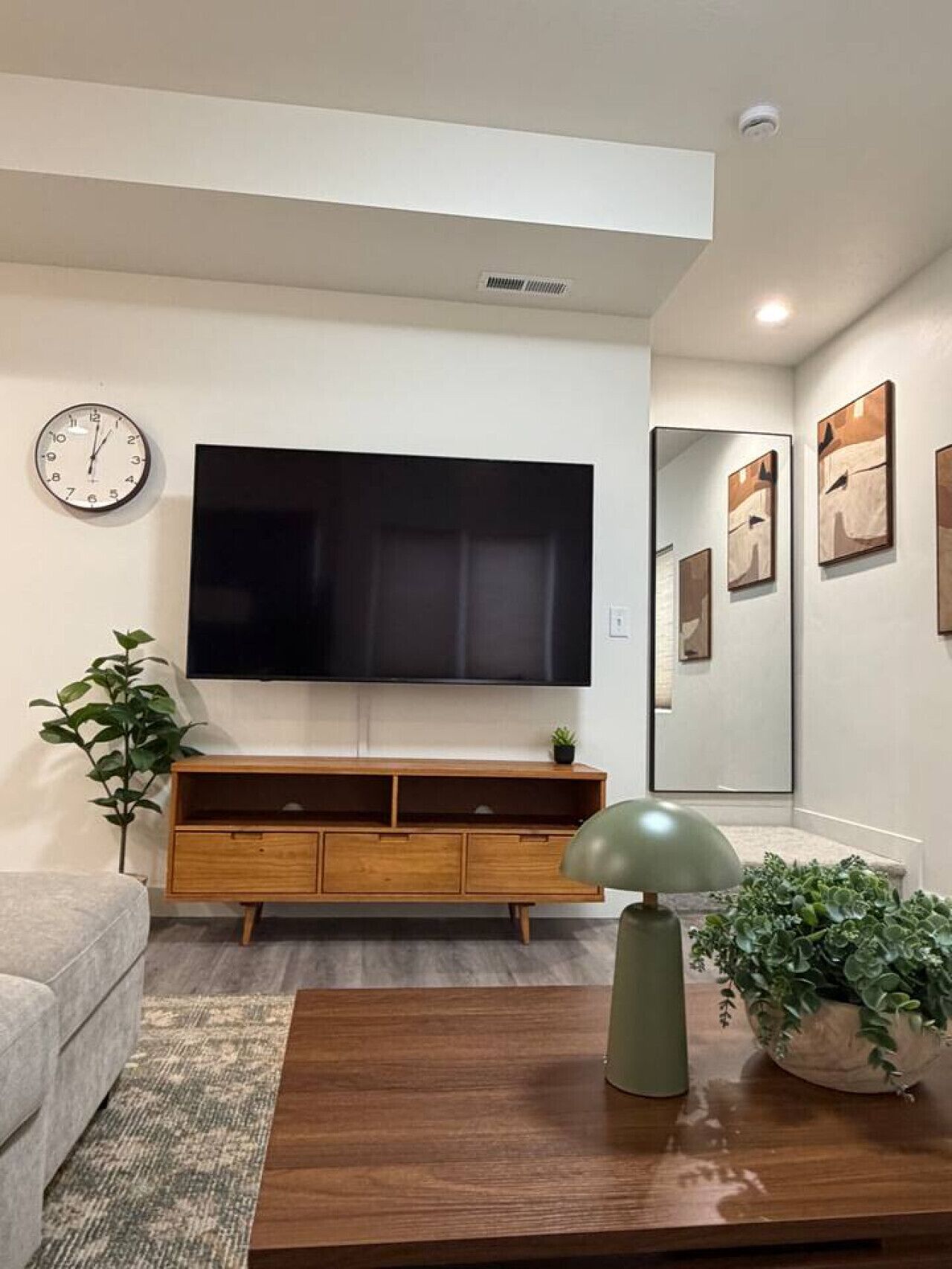 A living room with a flat-screen TV, wooden entertainment center, a clock, and a potted plant.