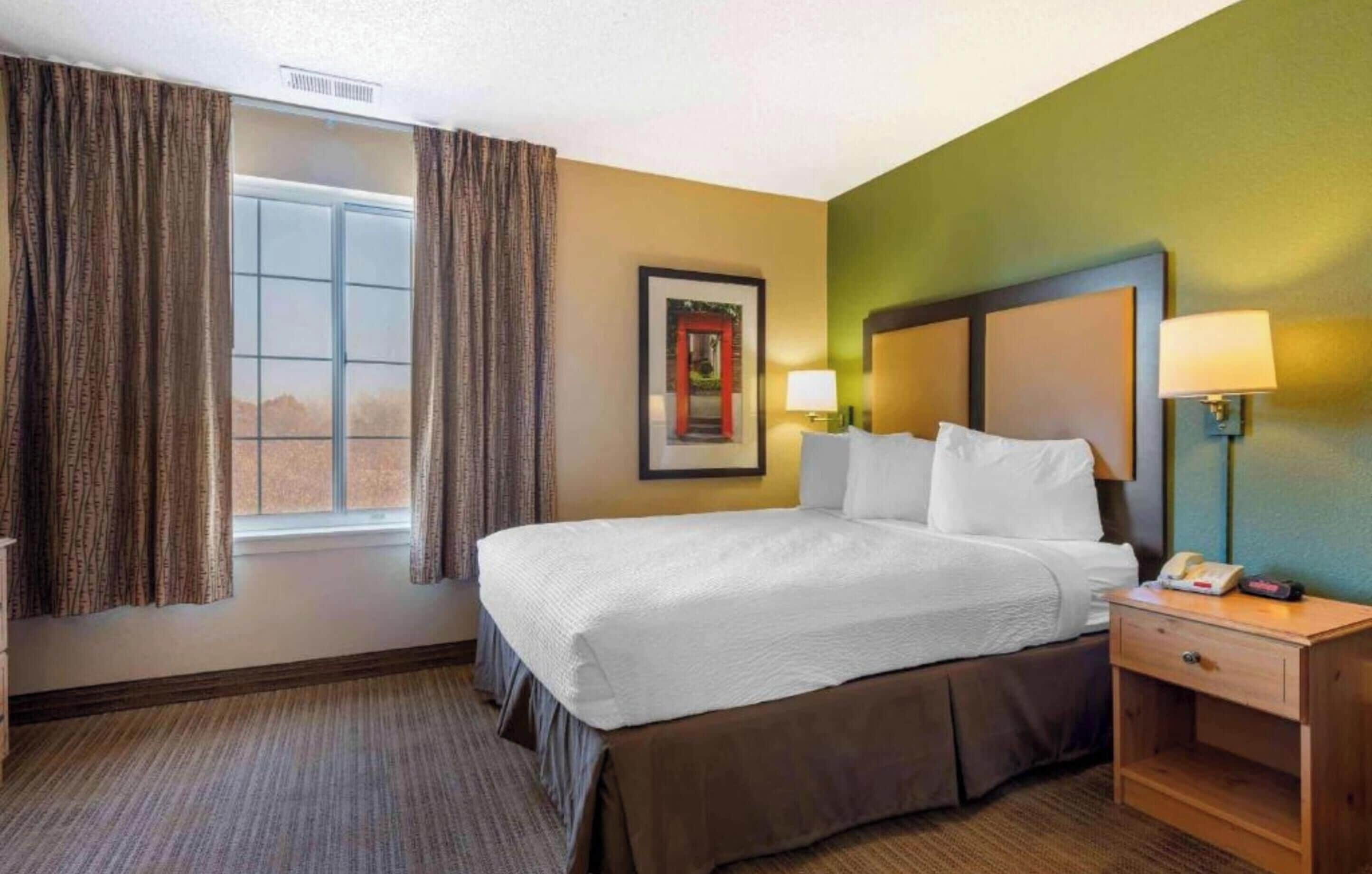 Habitación de hotel con cama, mesita de noche, lámpara, un cuadro enmarcado y ventana con cortinas.