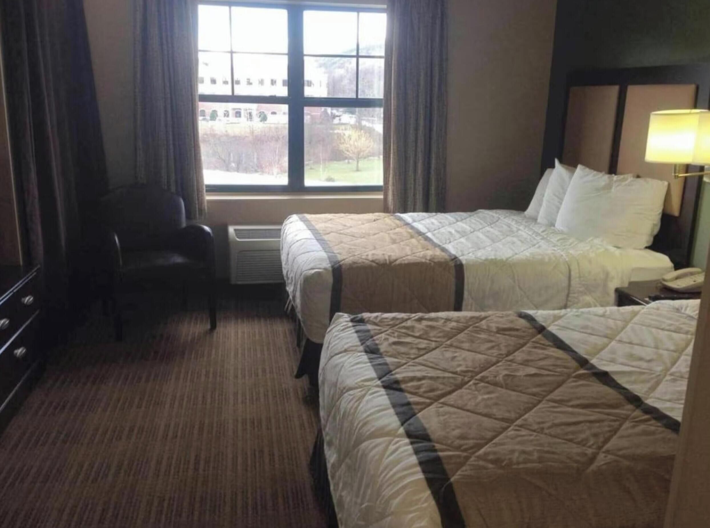 A hotel room with two beds, a chair, a desk, and a window with a view of a building and trees.