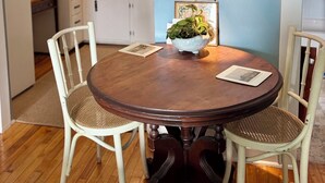 A dining area with a round wooden table, two chairs, a wall-mounted lamp, and a kitchen in the background.