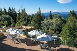 Ein möblierter Patio-Bereich mit mehreren Tischen und Sonnenschirmen, umgeben von dichtem Baumbestand und Blick auf ferne Berge.