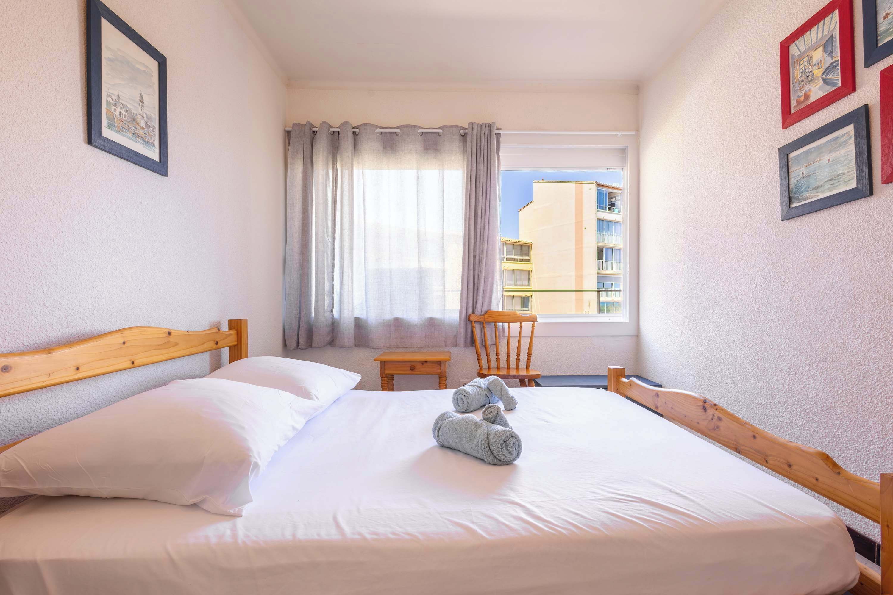 Un lit bien fait, avec du linge de lit blanc et des serviettes pliées, une tête de lit en bois, une petite table en bois et une chaise près d’une fenêtre ornée de rideaux.