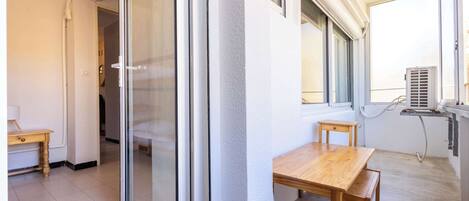 Una habitación con puerta corrediza de vidrio, una mesa de madera, un pequeño banco de madera y una ventana con unidad de aire acondicionado.