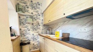 A compact kitchen with wooden cabinets, a marble backsplash, and a small trash bin. - Cozy Studio – Seaside Escape by the Water (Agde)