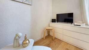 A modern living room with a flat-screen TV, a small stool, a white cabinet, and a decorative sculpture on a white table. - Cozy Studio – Seaside Escape by the Water (Agde)