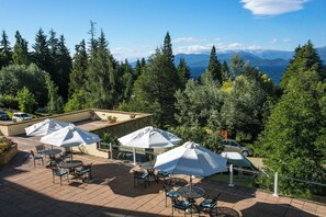 Eine Terrasse im Freien mit mehreren Tischen und Sonnenschirmen, umgeben von dichter Vegetation und Blick auf Berge und einen See.
