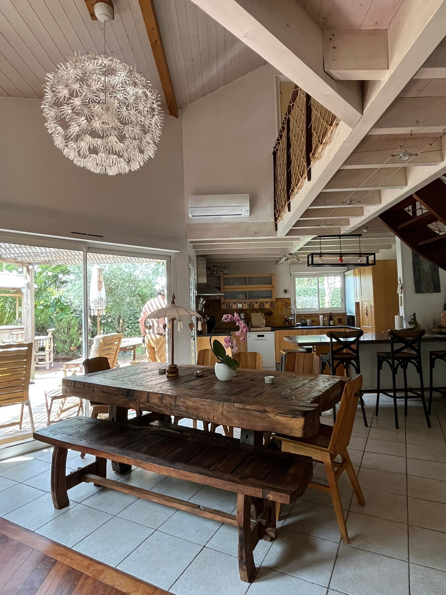 Un vaste espace de salle à manger avec une table et des chaises en bois, un luminaire suspendu et une vue sur la cuisine et l’espace extérieur.