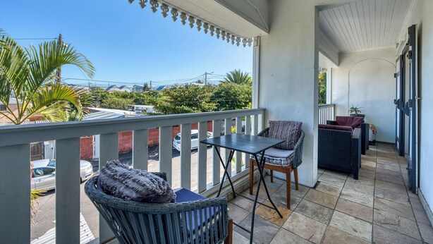 Ein überdachter Balkon mit Blick auf die Straße, ein kleiner Tisch, zwei Stühle und ein Korbsessel.