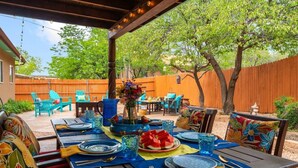Pátio externo com mesa de madeira preparada para refeições, rodeado de cadeiras e área de estar coberta.