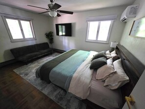 Ein Schlafzimmer mit Bett, Deckenventilator, Fernseher und zwei Fenstern.