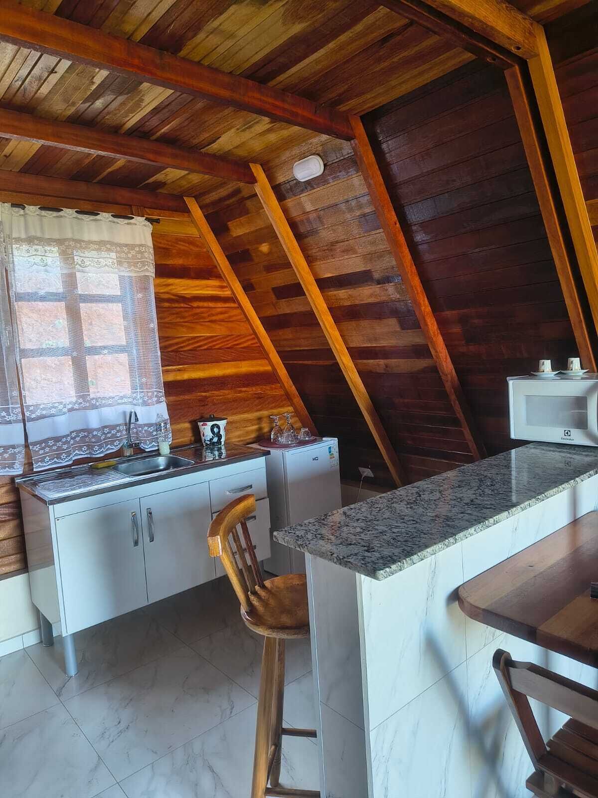 A kitchen with a wooden ceiling, a window with lace curtains, a sink, a refrigerator, and a bar stool.