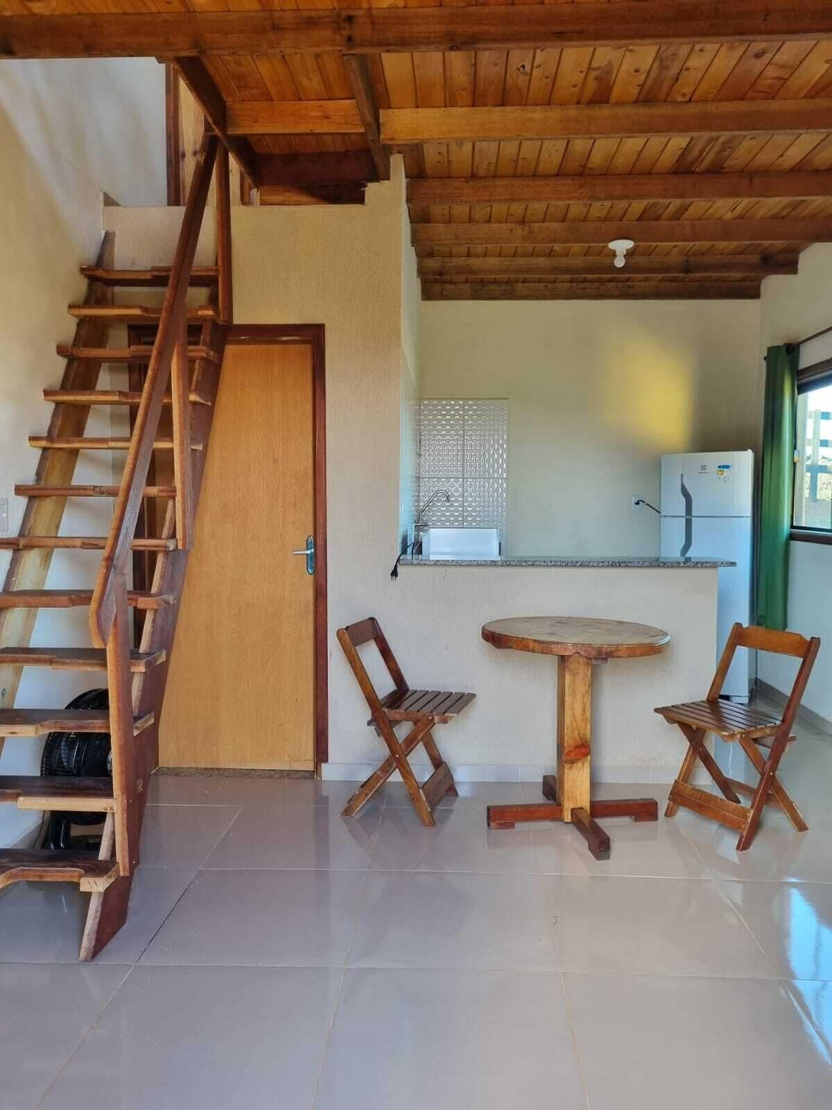 A kitchen with a refrigerator, a sink, and a window. There is a wooden staircase leading to a door. Two wooden chairs are placed around a small round table.